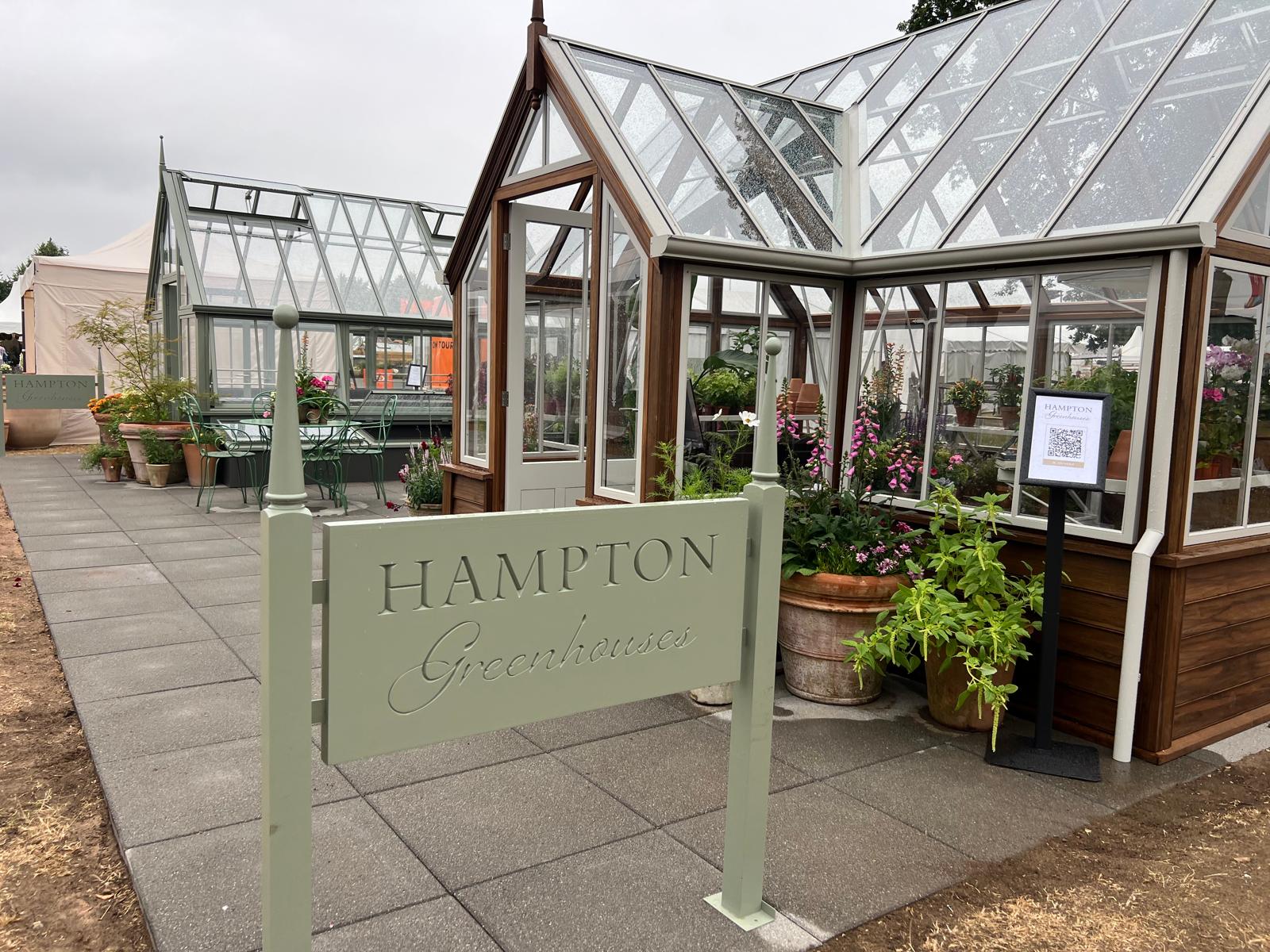 Two Hampton greenhouse models on display at the RHS show, with a wooden Hampton Greenhouses sign in front.