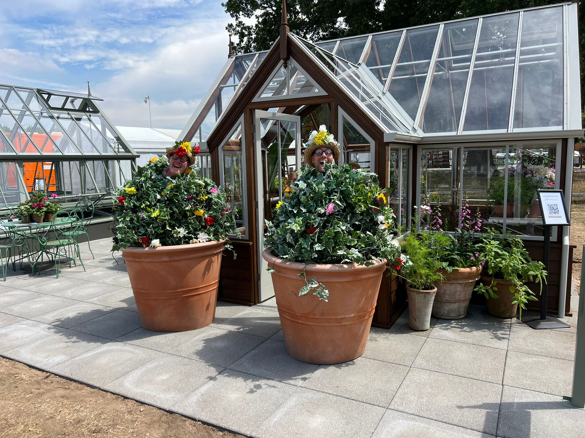 Two men in large flowerpot costumes in front of a Hampton greenhouse.