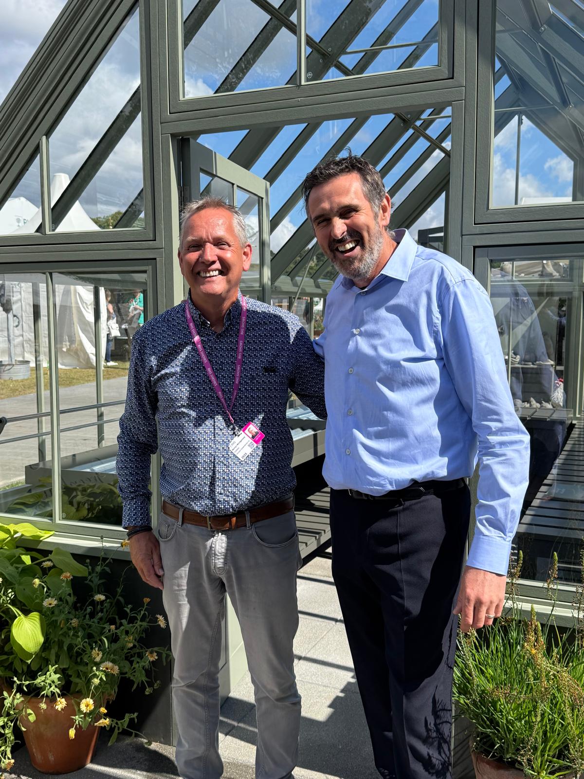 Mervyn Montgomery and Alan Frost, smiling together outside the front of a greenhouse.